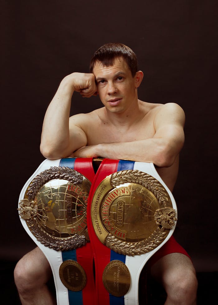 Portrait of a shirtless boxer sitting with championship belts, conveying victory and strength.